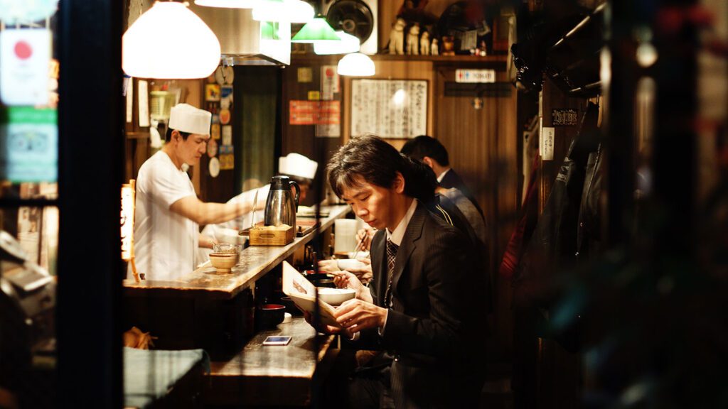 A businessman eating at a narrow, busy Japanese ramen bar while a chef prepares food in the background during his visit to Japan.