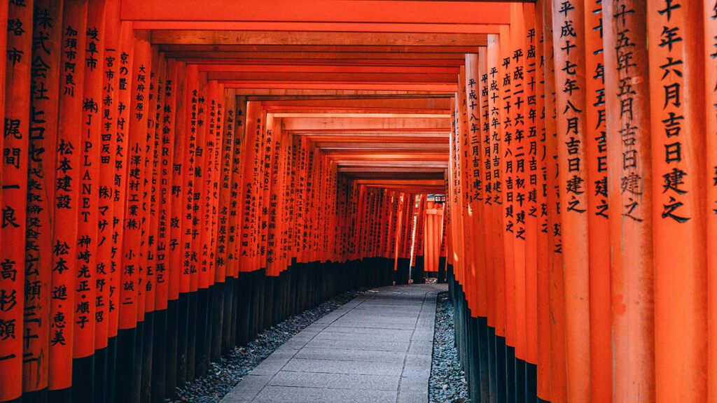 A pathway lined with vibrant red torii gates, each inscribed with Japanese characters, leading through a tranquil shrine invites visitors to explore one of the unique things to do in Japan.