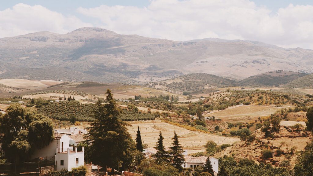 Scenic view of a lush valley in Spain with scattered trees and houses, backed by rolling hills and distant mountains under a clear sky.