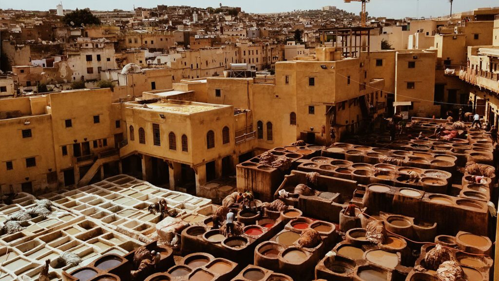 Aerial view of a traditional tannery with workers and dye-filled stone pits in a sunlit historic city in Morocco.