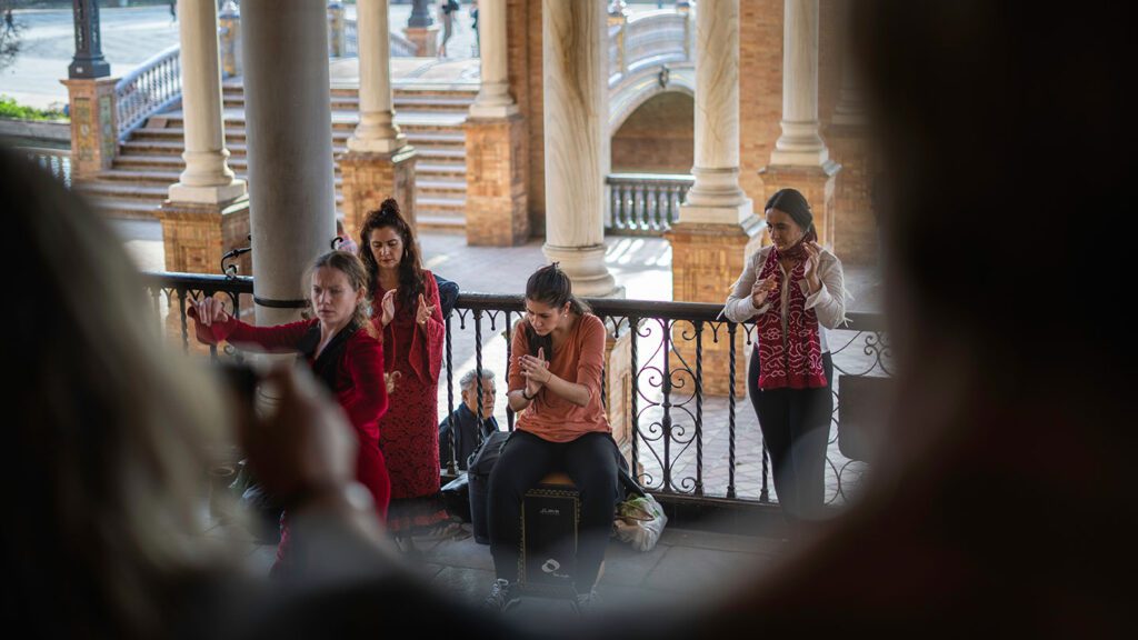 Group of people, mostly women, engaged in various activities (playing music, using a smartphone) on a balcony with ornate columns reminiscent of Morocco in the background.