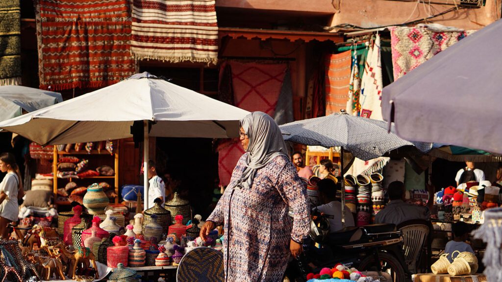 Woman in a hijab browsing a colorful market in Morocco with baskets and textiles under umbrellas in warm sunlight.