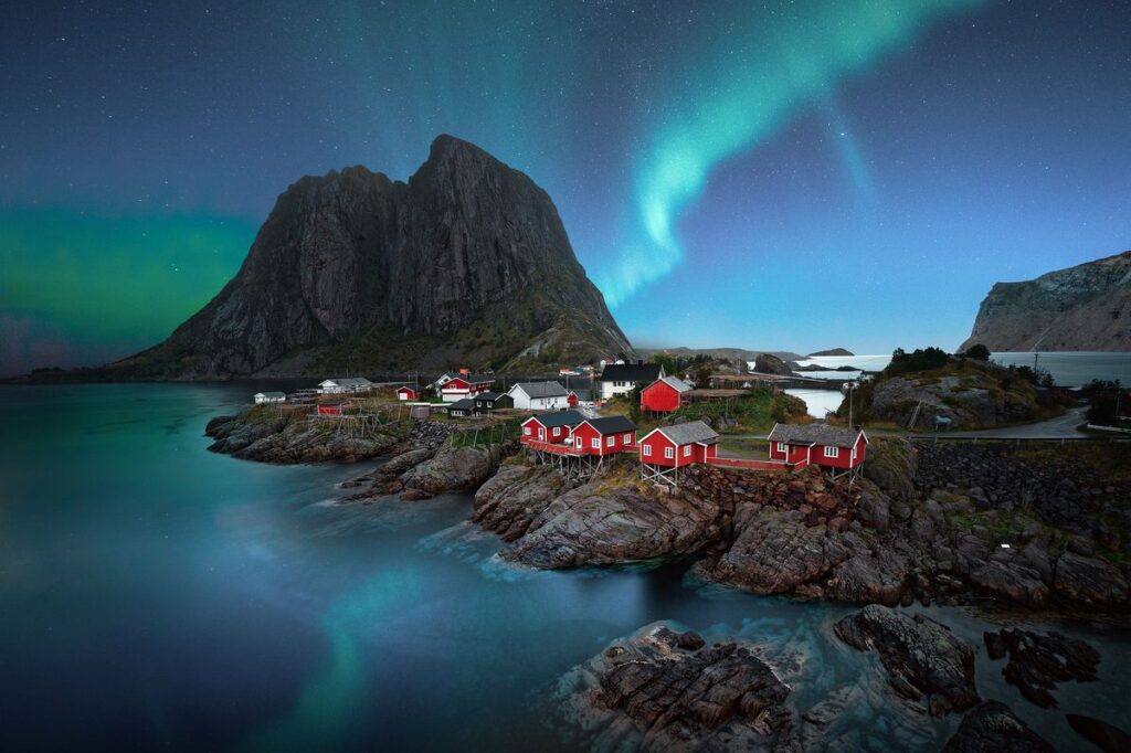 Norwegian village with red houses on a rocky peninsula under a night sky illuminated by the northern lights, a bucket list trip for 2023.
