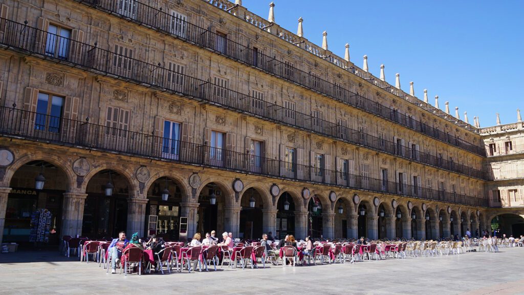 People dining at outdoor tables in Plaza Mayor, Salamanca, Spain, under the arches of a historic, ornately decorated building on a sunny day.