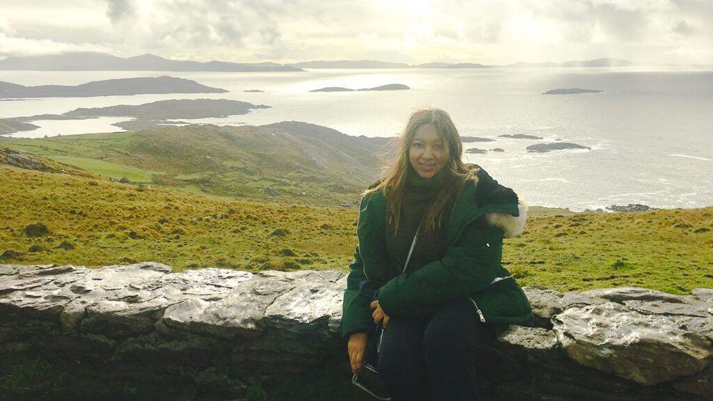 Woman sitting on a stone wall with a scenic view of a rugged coastline and islands in the background, under a cloudy sky.