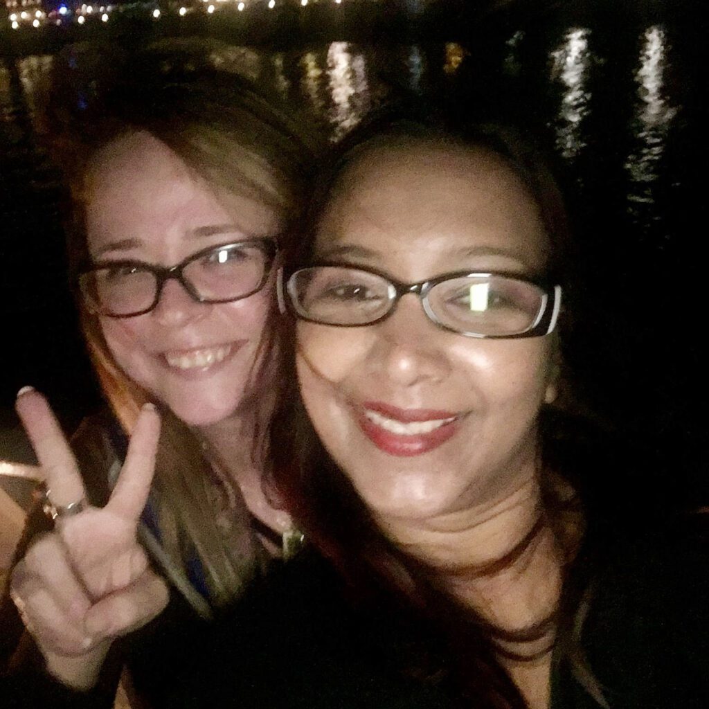 Two women smiling for a selfie at night, one making a peace sign, with lights reflecting on water in the background.