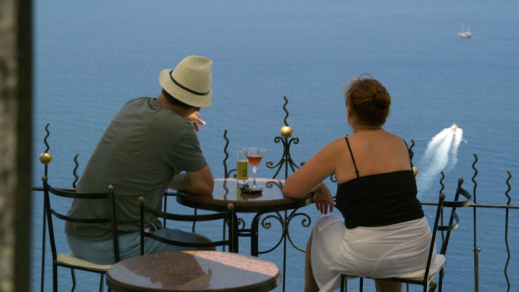A couple sits at a balcony table overlooking a lake, enjoying drinks and planning their next holiday, with a boat passing by in the distance.