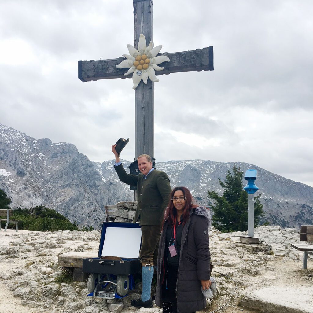 A man and a woman standing beside a large cross with a flower centerpiece on a mountain, with rocky peaks in the background.