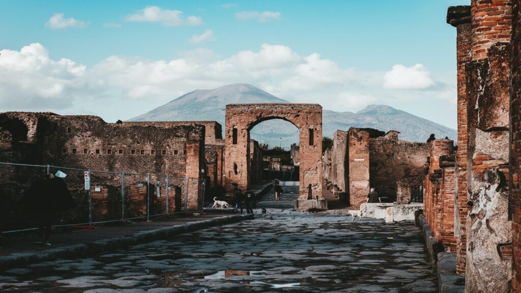 Ruins of ancient Pompeii with cobblestone paths, crumbling walls, and Mount Vesuvius in the background under a clear sky, a highlight on any Italy travel itinerary.