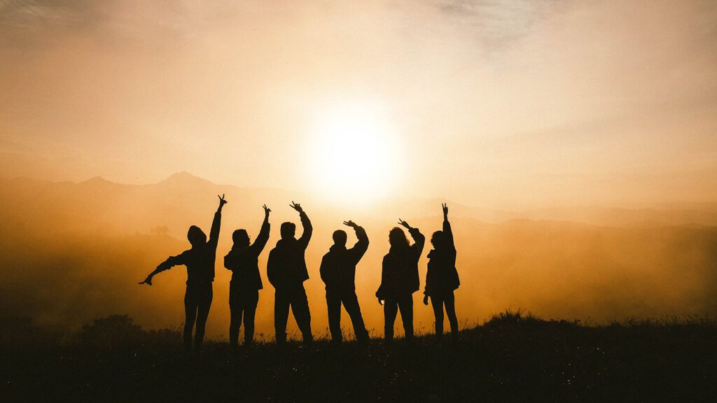 Silhouettes of seven people standing on a hill, raising their arms against a sunset background with mountains and a hazy sky, planning your next holiday.