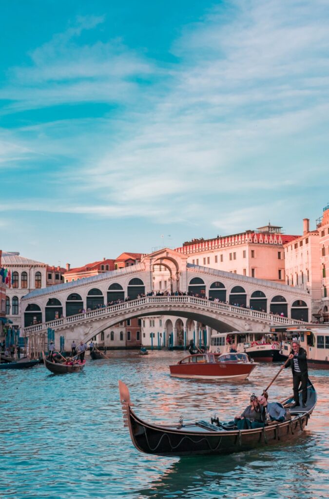 A gondola navigated by a gondolier on the Grand Canal in front of the Rialto Bridge in Venice, Italy, under a blue sky.