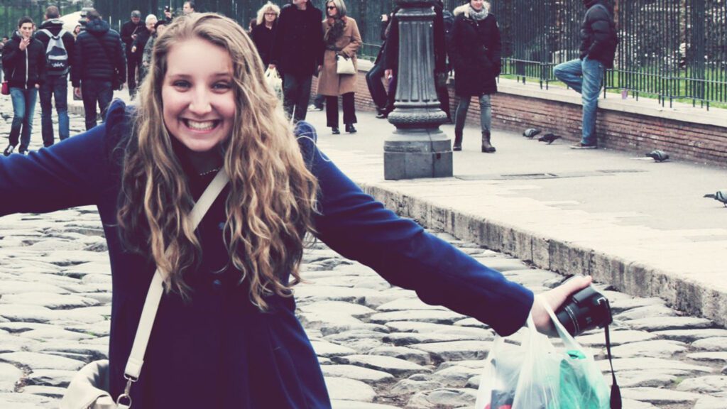 A young woman smiling joyfully with arms outstretched on a cobblestone street, carrying a camera and a shopping bag, surrounded by her best friends in the background.