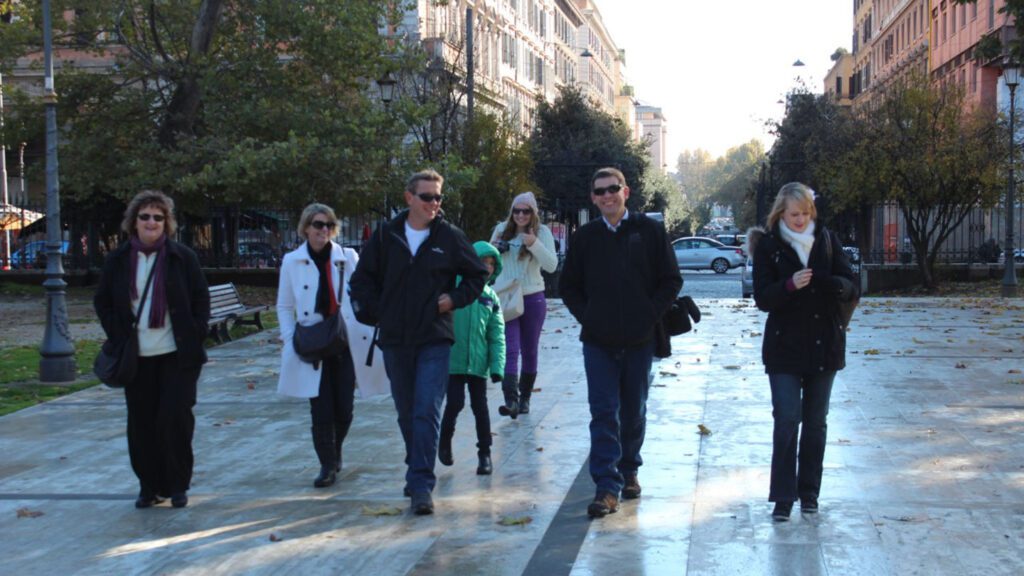 A group of seven best friends walking on a sunlit city street lined with trees, some are looking at their phones, others are talking.