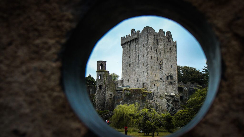 View of an ancient stone castle in Ireland, with a tower, framed within a circular hole, set against a backdrop of lush trees.