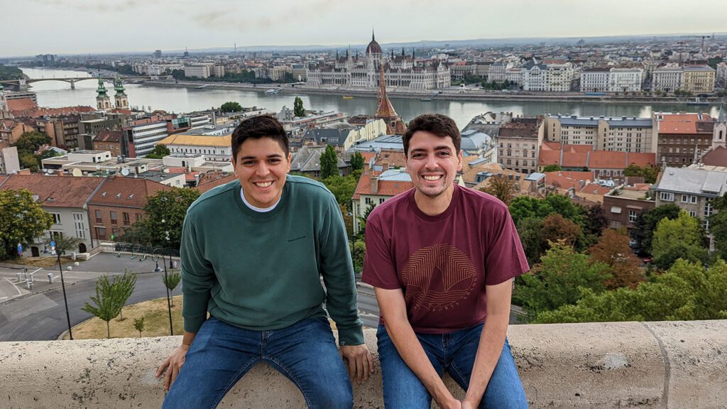 Two young men smiling at the camera while sitting on a wall, with a panoramic view of imperial Europe, including the Hungarian Parliament and a bridge over the Danube River, in the background.