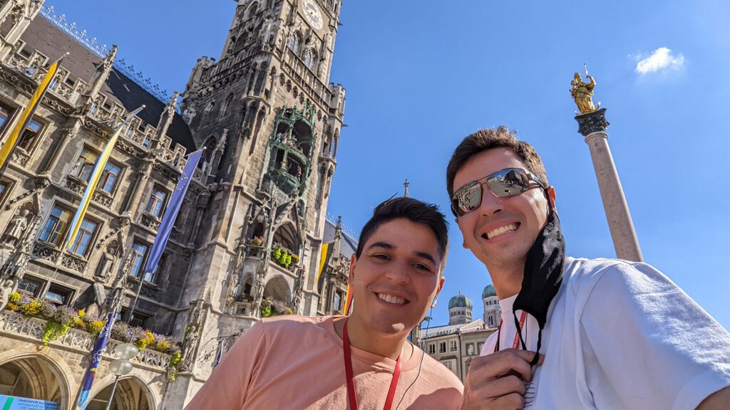 Two smiling men taking a selfie in front of the imperial new town hall and Marienplatz column in Munich on a sunny day.