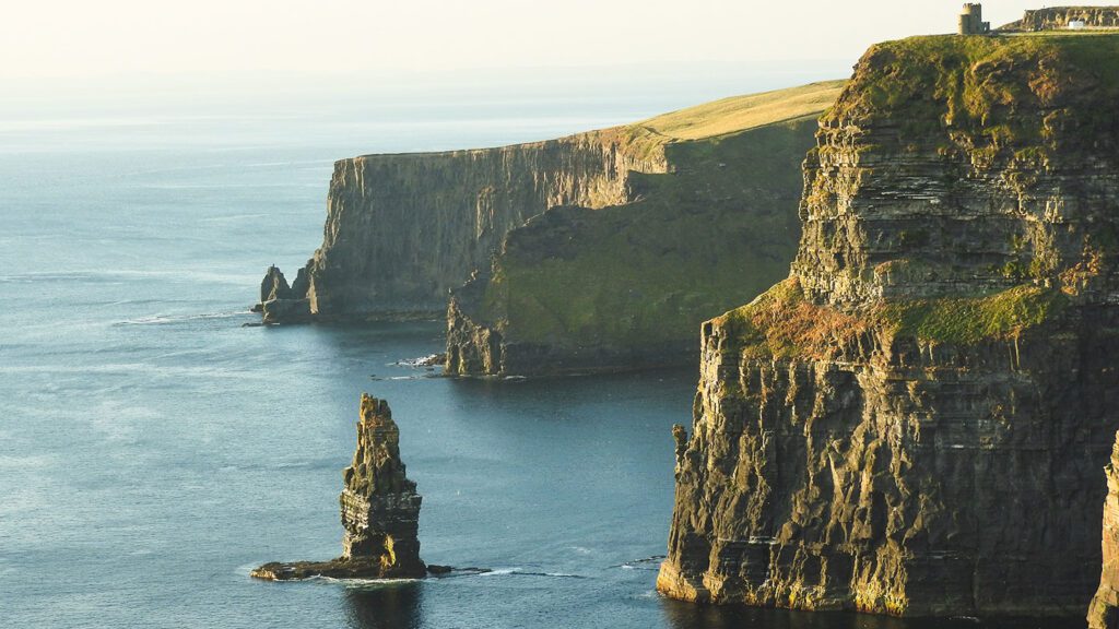 A panoramic view of the cliffs of Moher in Ireland, featuring tall, rugged cliffs beside a calm sea under a clear sky at sunset.