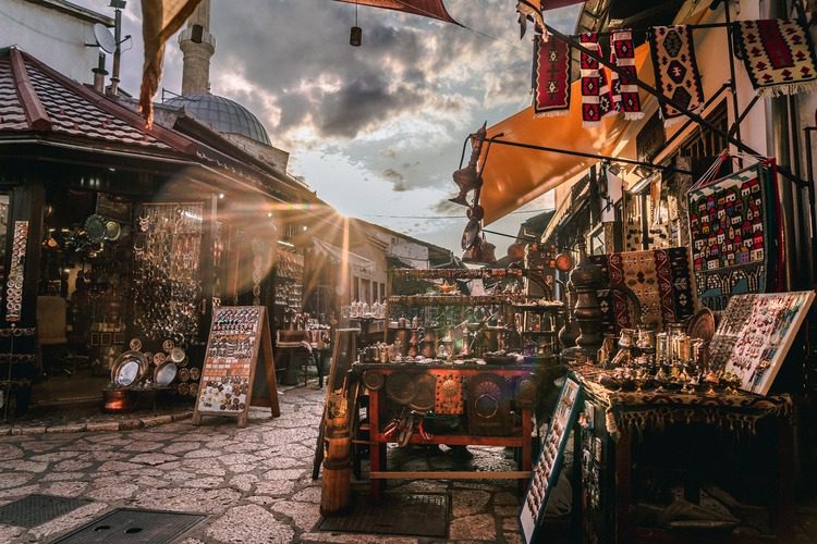Sunset over a bustling outdoor bazaar in Sarajevo with colorful rugs hanging, various merchandise stalls, and cobblestone streets leading to an old mosque.