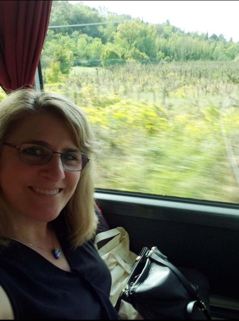 Woman with glasses smiling on a bus in Italy, with blurry green countryside visible through the window.