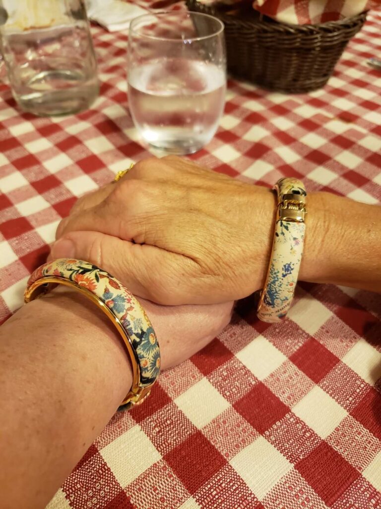 Two hands clasped together on a red and white checkered tablecloth, one hand adorned with floral pattern bracelets, reminisce about solo travelling in Italy.