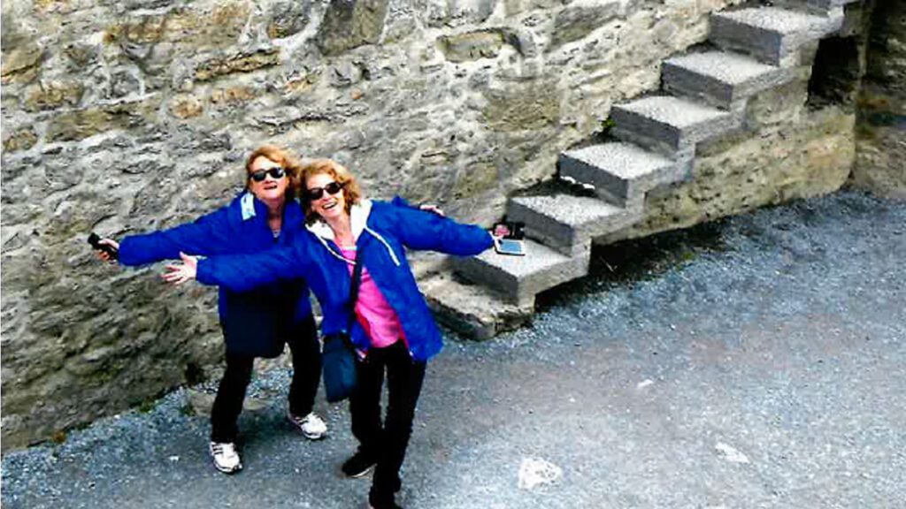 Two women with sunglasses, smiling and spreading their arms wide, standing near a stone stairway on a cobblestone path in Ireland.