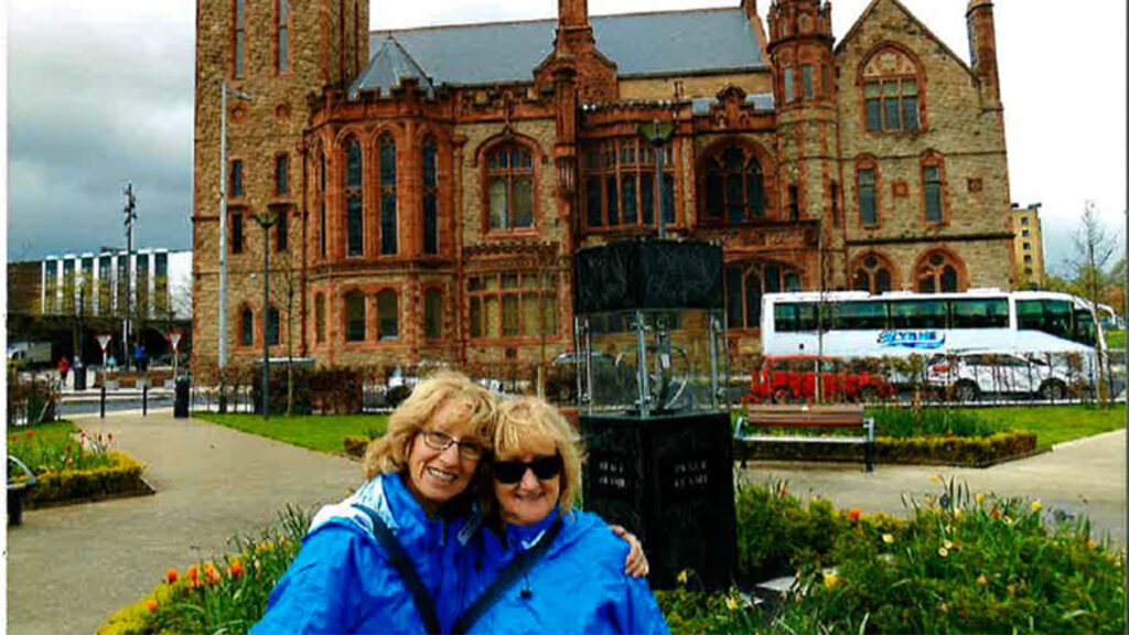 Two women in blue jackets smiling in front of a historic red brick building in Ireland, with a bus and greenery in the background.