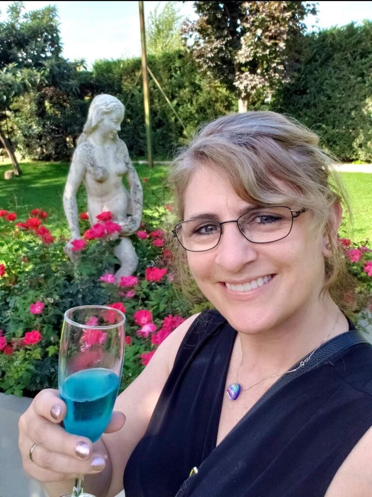 Woman smiling at camera holding a blue drink, with a statue and rose bushes in the background in a garden while solo travelling in Italy.