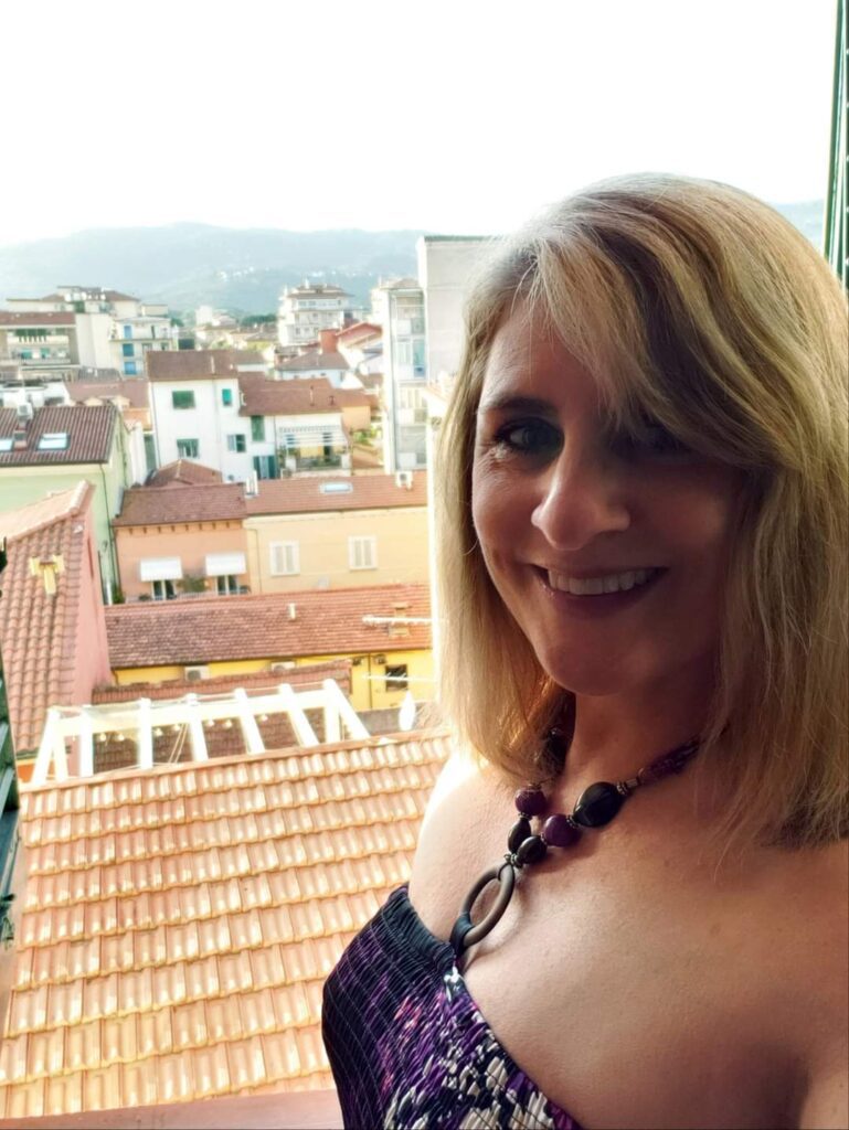 Smiling woman with shoulder-length blonde hair, solo travelling in Italy, standing on a balcony with city and mountain views in the background.