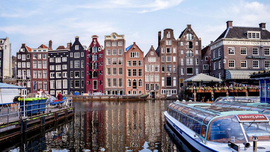 Traditional Dutch houses with gabled facades line the canal in Amsterdam, a popular summer destination in Europe, with tour boats moored along the water's edge.