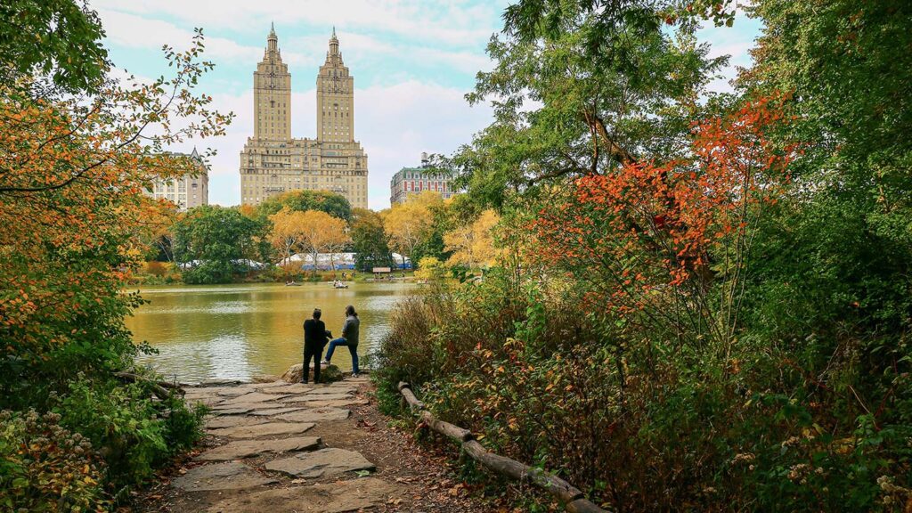 Two people by a lake in Central Park, overlooking towering buildings, surrounded by trees with autumn foliage, showcasing why it's one of the best cities to visit in September.