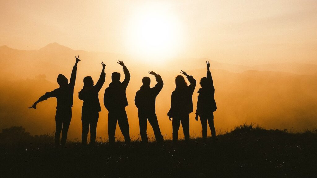 Silhouettes of five female travellers gesturing joyfully on a hill at sunset, with a hazy, golden sky in the background.