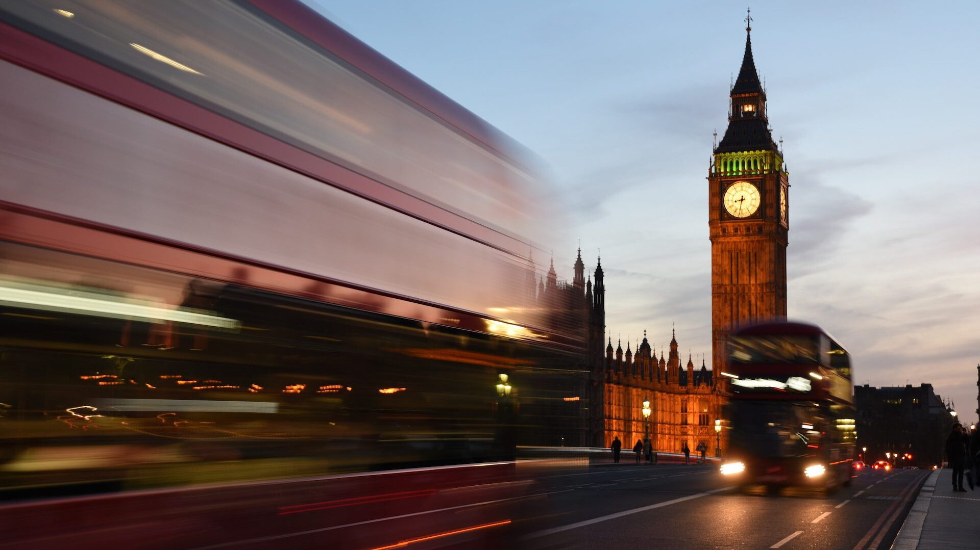 Image of London in the evening looking along Westminster Bridge towards Big Ben. Two double-decker buses in the foreground, blurred due to long exposure. 