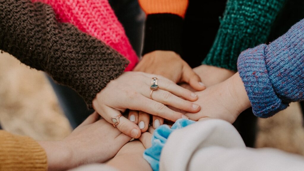 A group of female travellers join hands, displaying diverse knit sweaters and a visible engagement ring on a woman's hand.