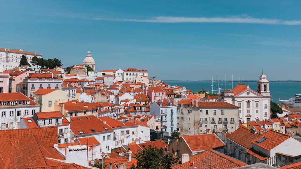 Panoramic view of Lisbon, one of the best European summer destinations, showcasing terracotta rooftops, white buildings, and notable landmarks under a clear blue sky.
