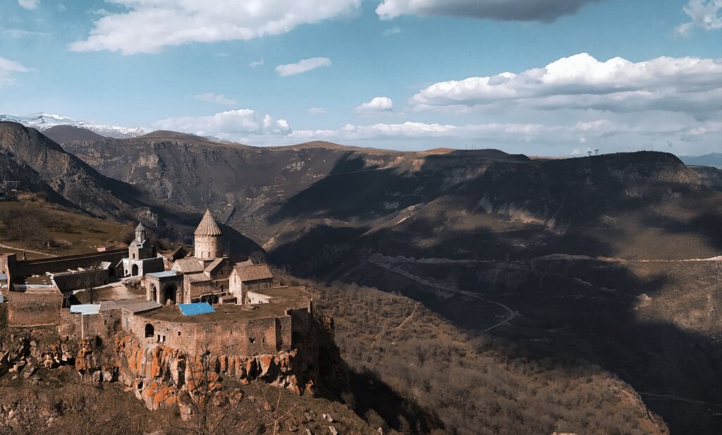 Ancient monastery with stone architecture perched on a cliff overlooking a mountainous landscape in Central & Eastern Europe with distant snow-capped peaks.