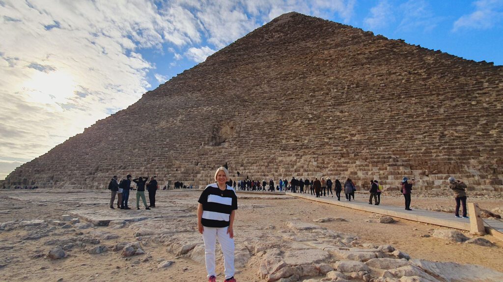 A solo female traveller standing in front of the great pyramid of Giza, with a clear sky and other visitors visible in the background.
