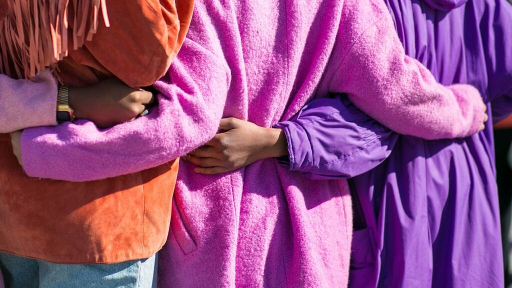 Three female travellers standing close together, each wearing brightly colored robes in orange, pink, and purple, with a focus on their linked arms.