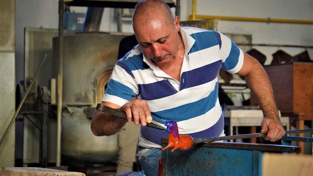 A glassblower molds a hot, molten glass piece into shape using a rod in his workshop, creating exquisite blown glass art.