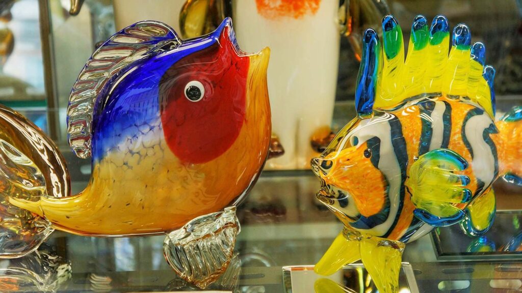 Colorful blown glass art sculptures of fish on display, featuring vibrant blue, red, and yellow tones with intricate details visible on a shelf.