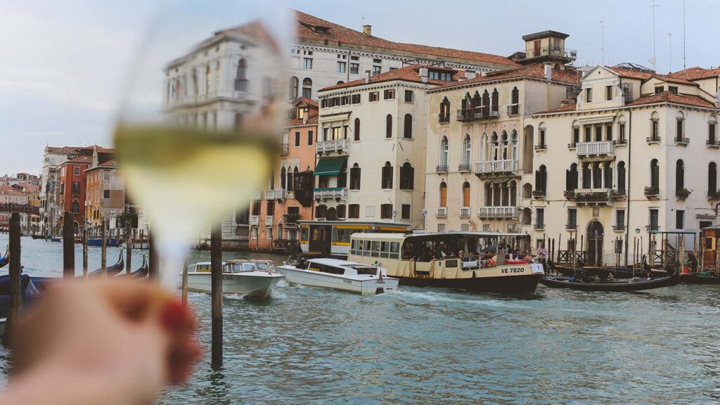 A close-up of a hand holding a glass of Venetian glass white wine with a blurry backdrop of the Grand Canal in Venice, showing boats and historical buildings.