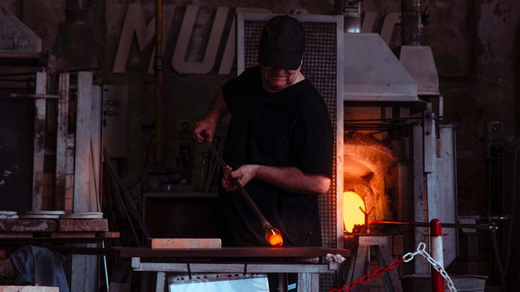 A glassblower in a dark workshop shapes a molten Venetian glass piece using a metal rod, with a glowing furnace nearby.