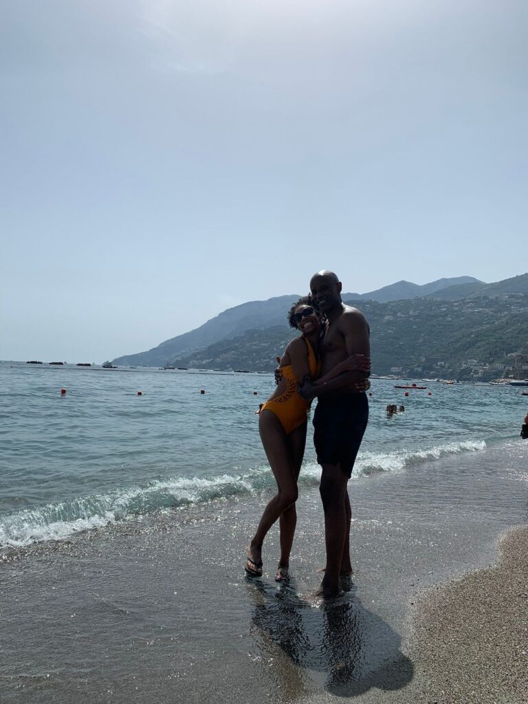 A couple embracing on a pebbly beach with waves at their feet and a hazy mountain landscape in the background, capturing one of their honeymoon stories.