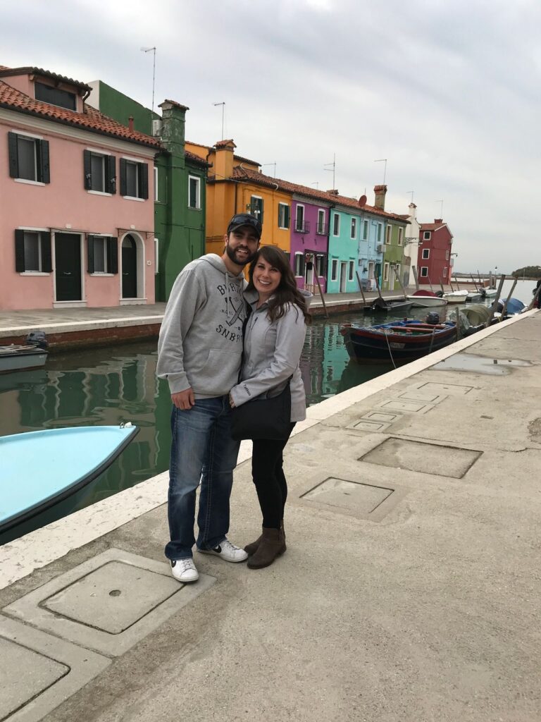 A couple smiling and posing together in front of a canal with colorful houses and boats in Burano, Italy, capturing honeymoon stories.