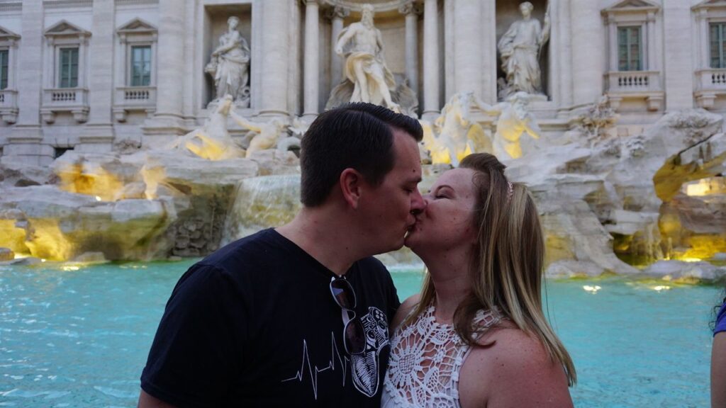 A couple kissing in front of the Trevi Fountain in Rome, Italy, with statues and flowing water visible in the background, capturing a moment from their honeymoon stories.
