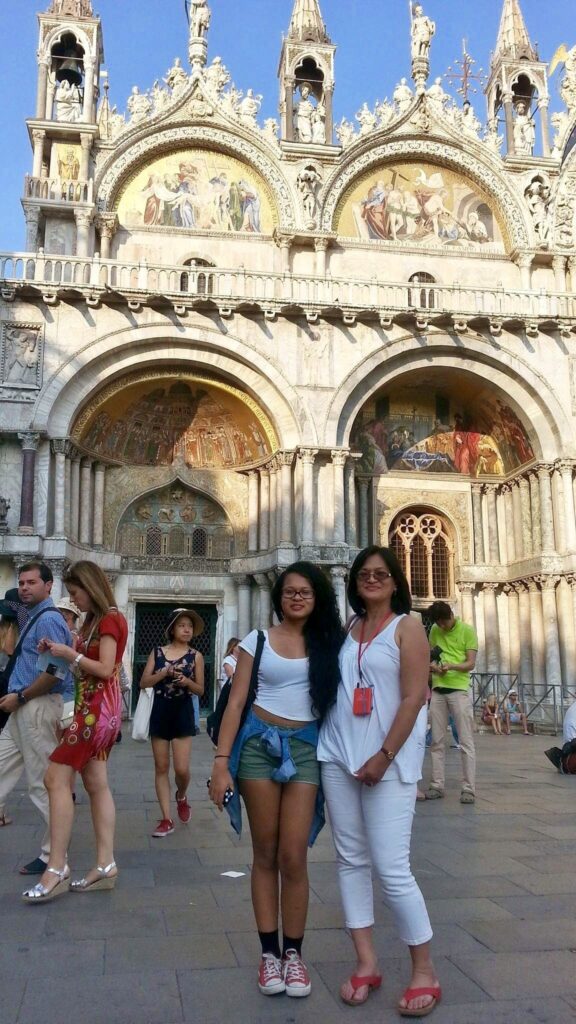 Two women posing in front of Saint Mark's Basilica in Venice, Italy, celebrating Mother's Day, surrounded by other tourists.