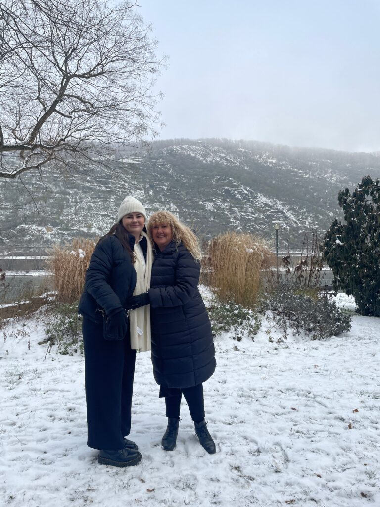 Two women smiling and hugging each other in a snow-covered park with bare trees and a hill in the background on Mother's Day.