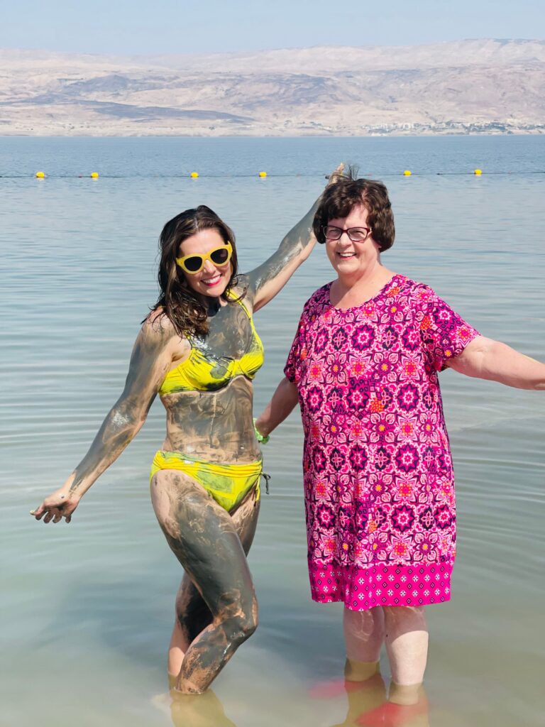 Two women, one covered in mud and wearing a bikini, and the other in a pink floral dress, celebrating Mother's Day by standing in a lake, smiling.