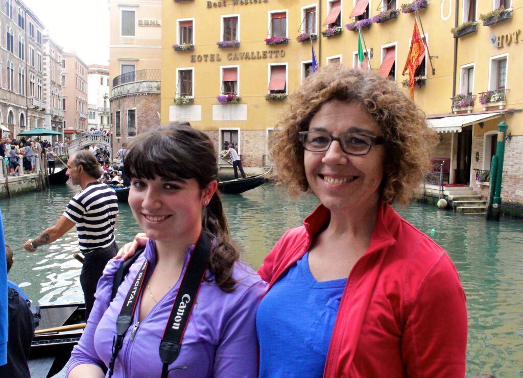 Two women smiling beside a canal on Mother's Day, with a gondola and gondolier in the background, colorful buildings and a hotel sign visible.