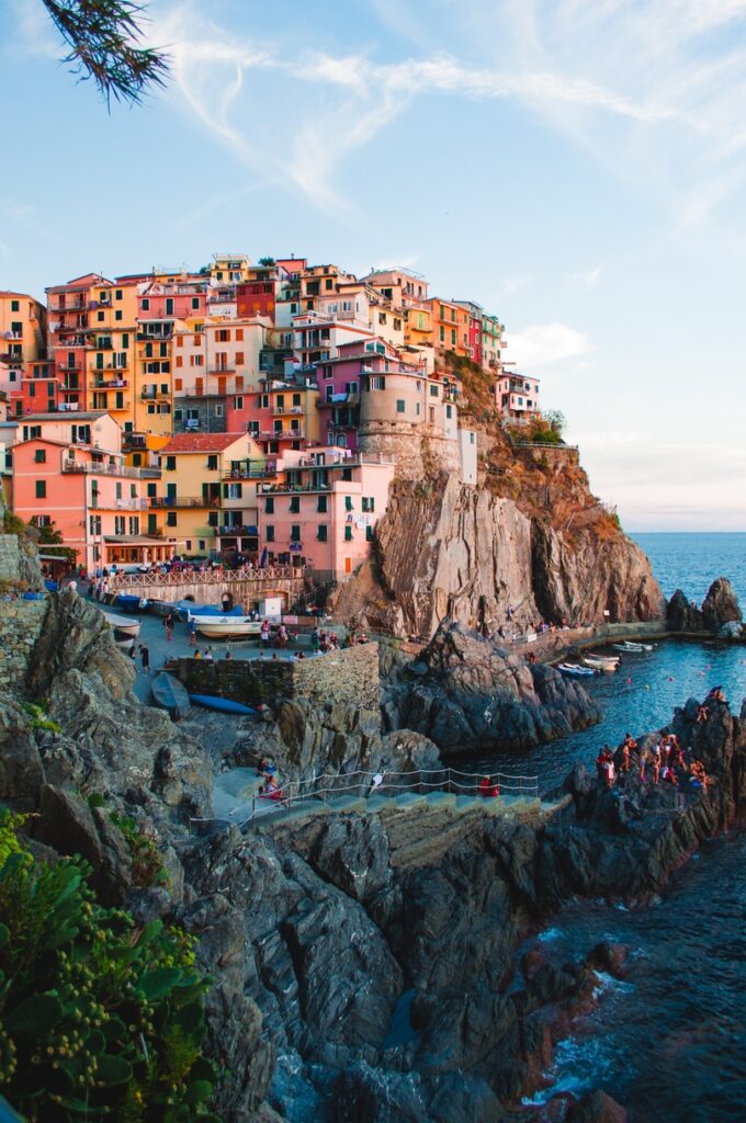 Colorful buildings perched on a rugged cliff overlooking the sea, with tourists gathered by the shore at sunset in Cinque Terre, Italy.