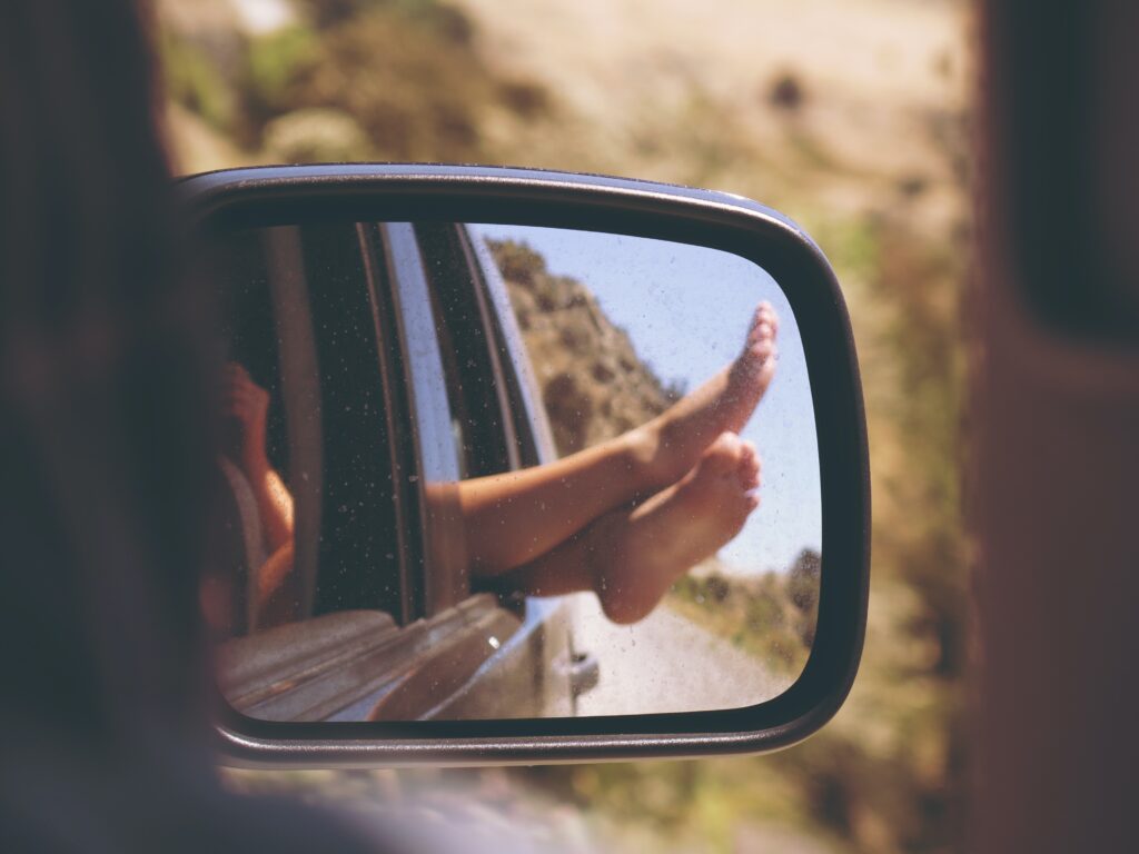 A car's side mirror reflects a child's bare feet resting on the window frame, with a blurred landscape in the background, keeping kids entertained during the drive.